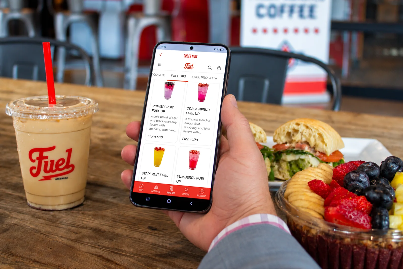 Fuel America app on a smartphone beside an iced coffee and café food